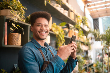 Cheerful florist with a big smile typing on smartphone in flower shop. Happy flower shop worker in a brown apron using mobile phone surrounded by vibrant plants and fresh flowers