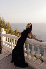 beautiful woman with dark hair in luxurious black dress with accessories posing at the balcony of villa with sea view