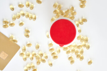 Top view of vitamin capsules scattered around a red-lidded container and bank card on white background