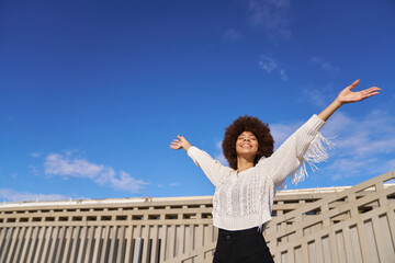 A woman with curly hair is standing outside with her arms open, looking up at the sky