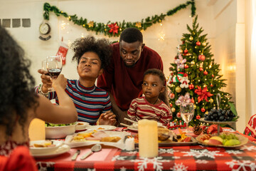 A joyful family gathers around the dinner table celebrating Christmas. The father, dressed in a Santa hat, gives a gift to the child, while everyone smiles with warmth and festive decorations.