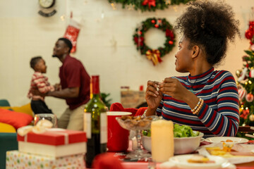 A joyful family gathers around the dinner table celebrating Christmas. The father, dressed in a Santa hat, gives a gift to the child, while everyone smiles with warmth and festive decorations.