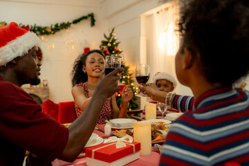 A joyful family gathers around the dinner table celebrating Christmas. The father, dressed in a Santa hat, gives a gift to the child, while everyone smiles with warmth and festive decorations.