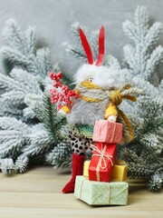 Christmas character rabbit with colorful ears in a sweater with presents in his hands on the background of pine branches