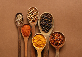 Mix of spices in wooden spoon isolated on a brown background. Top view. Flat lay. Set or collection of condiments of black pepper, curcuma, paprika, cumin, coriander seeds and chilly. 