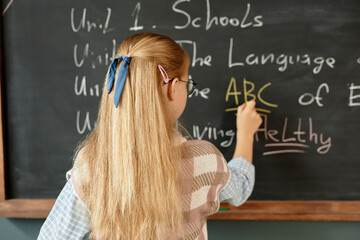 Back view of young girl with long blonde hair decorated with ribbon writing letters on blackboard...