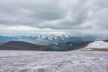 Dramatic panoramic view from big glacier to wide alpine valley and large snow-capped mountain range in rainy low clouds. Awesome vast landscape with high snowy mountains in rain under gray cloudy sky.