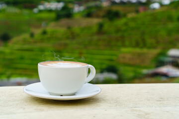 White cup of hot latte art coffee on cream wooden table with terrace mountain background in rainy season.