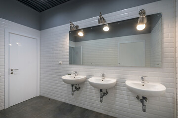Office public bathroom, decorated with white tiles. A large mirror and three sinks.