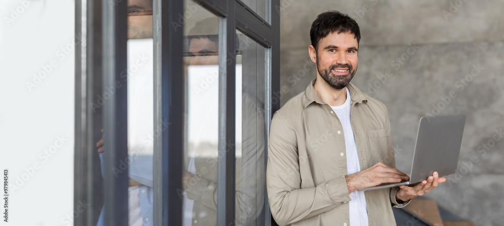 Poster A man stands smiling while using a laptop near a large window in a contemporary office. The natural light enhances the workspace, creating a productive atmosphere.