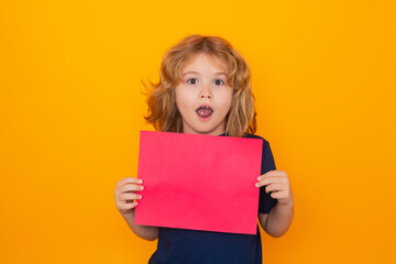 Kid with empty sheet of paper, isolated on yellow background. Portrait of a kid holding a blank placard, poster. Copyspace. Surprised face, amazed emotions of child.