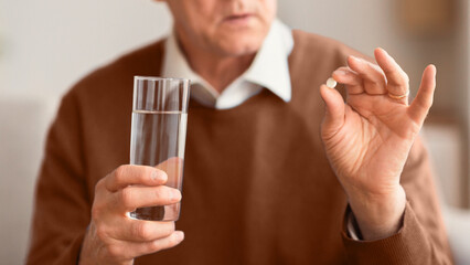 Unrecognizable Elderly Gentleman Holding A Pill And Glass Of Water Sitting On Couch Indoor. Cropped, Selective Focus