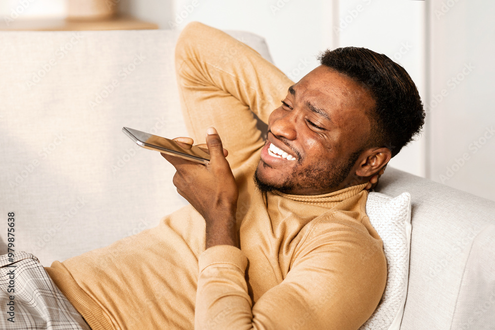 Poster modern lifestyle. happy afro man using phone loudspeaker or voice assistant lying on sofa at home.