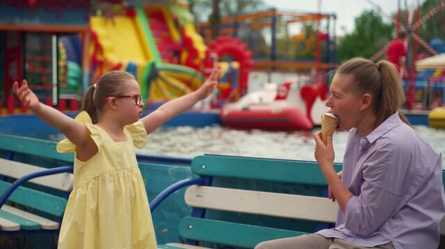Mother enjoys ice cream while her young daughter with Down syndrome performs a joyful dance at amusement park. Scene captures happiness and playful moments of nurturing child with special needs