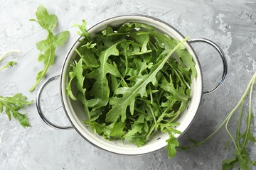 Fresh green arugula leaves in colander on grey textured table, top view