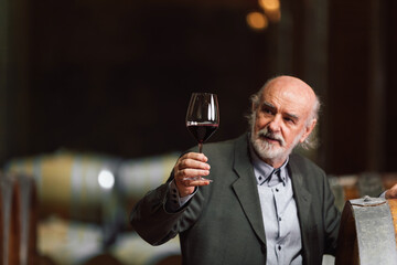 Caucasian senior man in a suit, standing in an old wine cellar with wooden barrels, expertly tasting red wine, checking a color, smell, and taste