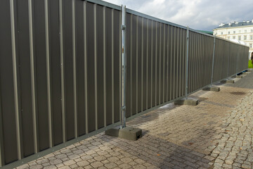 A modern gray fence creates a barrier in an urban setting, casting shadows on the cobblestone pavement below. Ideal for construction or safety themes.