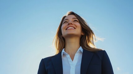 A businesswoman confidently gazes upward while standing outdoors. She wears a tailored suit and smiles, exuding positivity beneath a clear blue sky