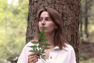 young woman in the forest near a tree holds a fern in her hand