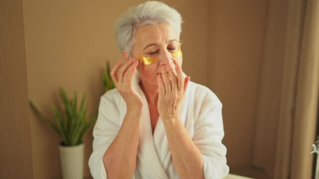 Eye patch beauty aging treatment. Mature adult old woman with under eye collagen pads. Senior woman with patches under eyes gets facial treatment. Senior lady in robe relaxing in bathroom at home
