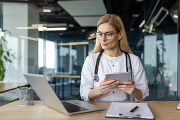 Healthcare professional wearing glasses and stethoscope uses tablet and laptop in modern office for research and communication. Focused medical expert demonstrating technology use in healthcare