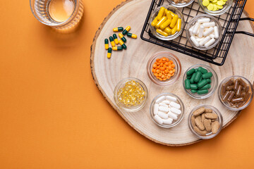 Colored pills and tablets of dietary supplements in small glass bowls and a glass of water on a wooden desk from above on an orange background. Groups of vitamins and minerals top view. Rustic style.