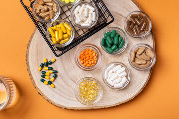 Colored pills and tablets of dietary supplements in small glass bowls and a glass of water on a wooden desk from above on an orange background. Groups of vitamins and minerals top view. Rustic style.