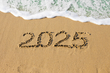 Fototapeta premium 2025 new year concept. Text 2025 handwritten on sand surface. Frothy wave washing away text on a sand beach.