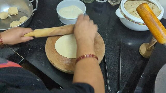In the kitchen, closeup of a womans hand rolling out round dough to prepare round flatbread roti using a wooden roller and board