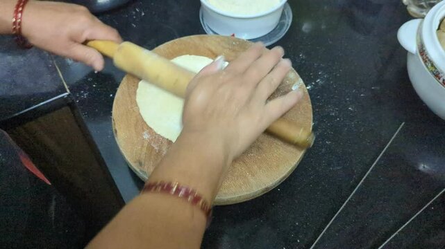 In the kitchen, closeup of a womans hand rolling out round dough to prepare round flatbread roti using a wooden roller and board