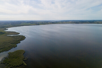 Aerial view of a tranquil lake surrounded by greenery, reflecting a clear sky. The landscape captures serenity and natural beauty