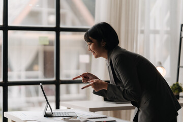 Engaged in the Digital Dialogue: A young Asian businesswoman, bathed in warm light, leans in with a bright smile as she engages in a video conference on her laptop.