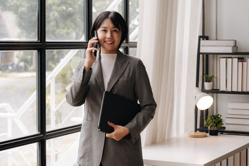 Confident Businesswoman on the Phone: A professional woman in a gray blazer smiles confidently while on a phone call, holding a black folder in her hand. She stands near a large window in a bright off