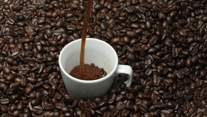 Freshly espresso was poured in white cup with pile of coffee bean. Pouring black coffee in the cup with brown background and seed scattering around on wooden table. Close up. Top view. Comestible.