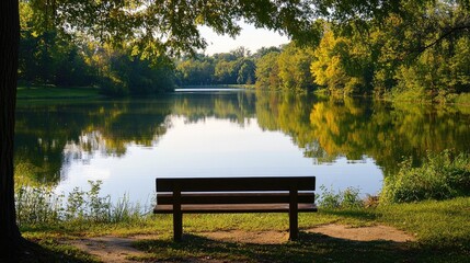 Obraz premium Quiet park bench by a tranquil lake with reflections of surrounding trees and a clear sky.