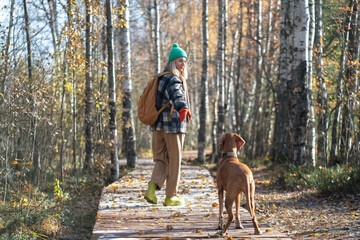 Smiling woman strolling along wooden path through dense woods with pet, rear view. Female traveler...