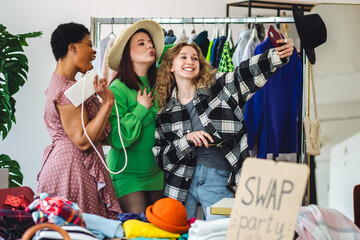 Cheerful young student friends having fun at home swap party, exchanging clothes, trying hats. Eco...