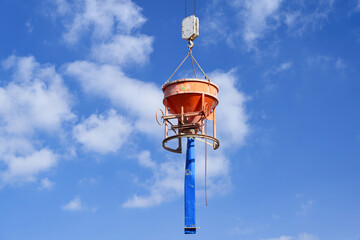An orange concrete bucket with a blue hose for transporting fresh concrete hangs on a crane hook on a construction site against a blue sky
