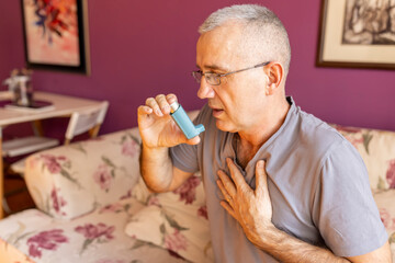 Senior man sitting on sofa at home and holding hand on chest. Male having asthma attack,...