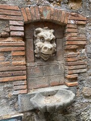 Ancient stone fountain with grotesque gargoyle head mounted in brick-framed niche. Weathered stone basin below. Medieval architectural detail showing historic craftsmanship and European character.