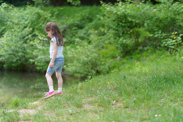 Little cute preschool girl in a T-shirt plays on the background of a pond. Happy childhood concept outdoors