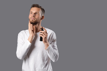 Handsome young man applying cosmetics for beard on grey background