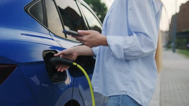 Female driver of eco-friendly car pays online for service via cellphone. Woman with smartphone plugs out electric vehicle at charging station