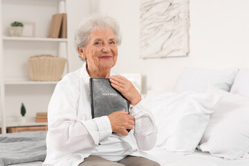 Senior woman with Bible sitting on bed at home