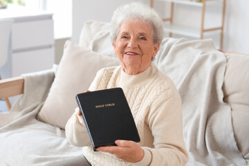 Senior woman with Bible sitting on sofa at home