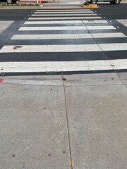 A clear midday view of a classic black-and-white striped crosswalk in Santa Monica, California. The sun casts minimal shadows, highlighting the textures of the worn crosswalk.
