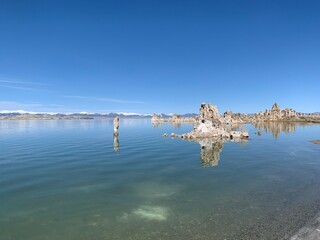 A wide shot of the calm water surface of Mono Lake on a clear day, revealing the slightly visible lakebed near the shoreline. In the background, snow-capped mountains stand tall on the horizon.