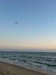 A serene beach scene with a commercial airplane soaring overhead, captured at Venice Beach, Los Angeles, with the vast Pacific Ocean stretching out to the horizon. The tranquil atmosphere.