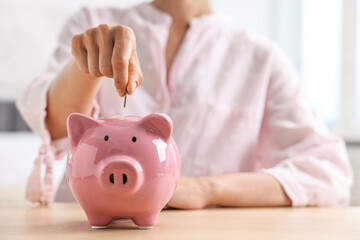 Woman putting coin into piggy bank at table indoors, closeup