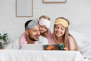 Happy parents and their little daughter with sleeping masks reading book in bedroom